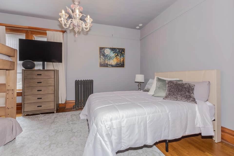 Bedroom with white bed, wooden dresser, TV, pale purple walls, hardwood floors, and crystal chandelier