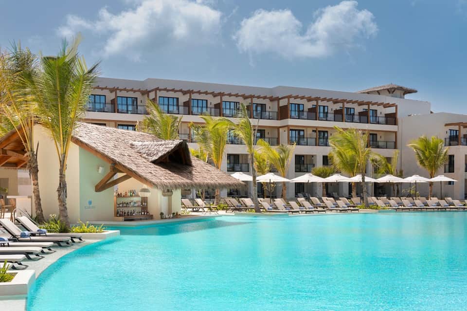 Tropical resort pool with thatched-roof bar, palm trees, white umbrellas, and loungers