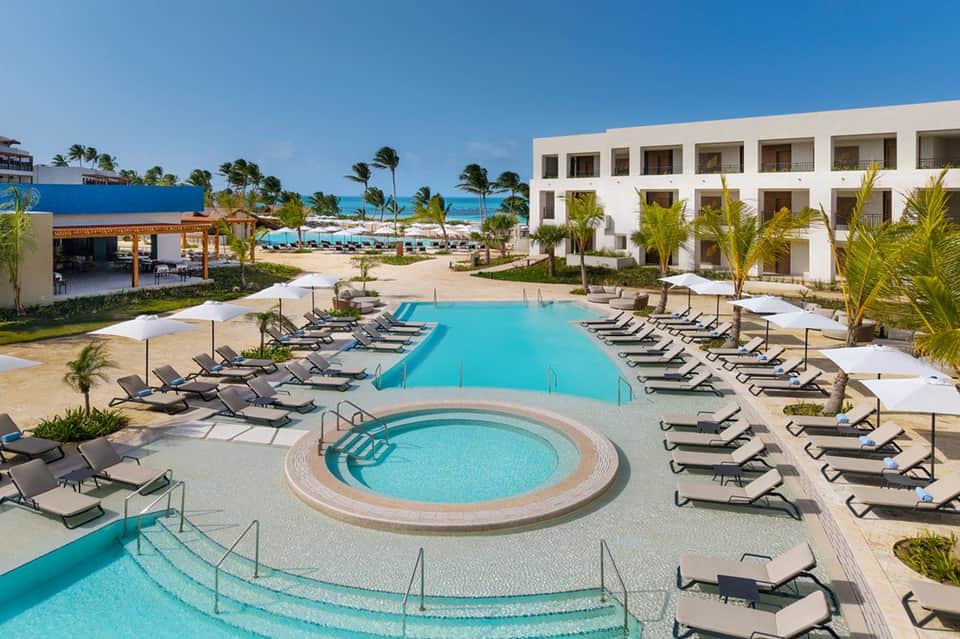Resort courtyard with turquoise pool, spa tub, white building, and ocean view in background