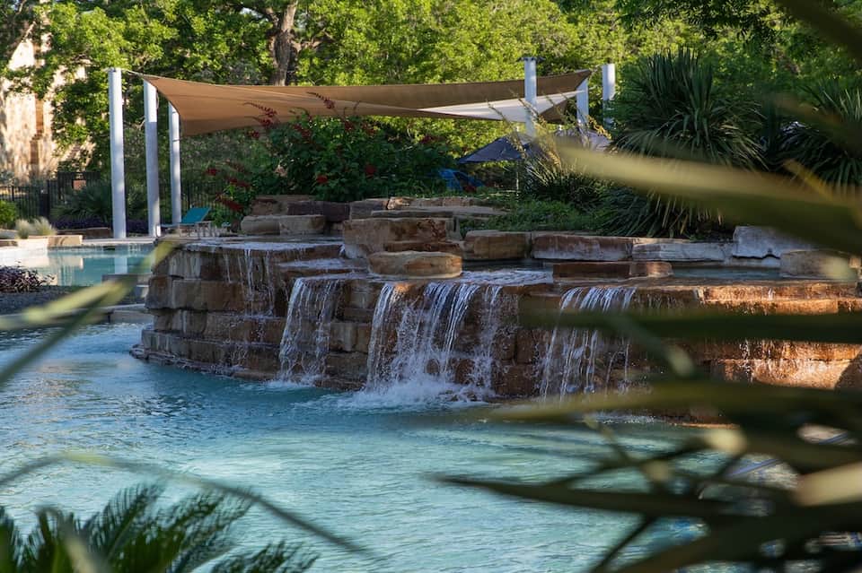 Resort swimming pool with waterfall feature and sail shade structure surrounded by trees