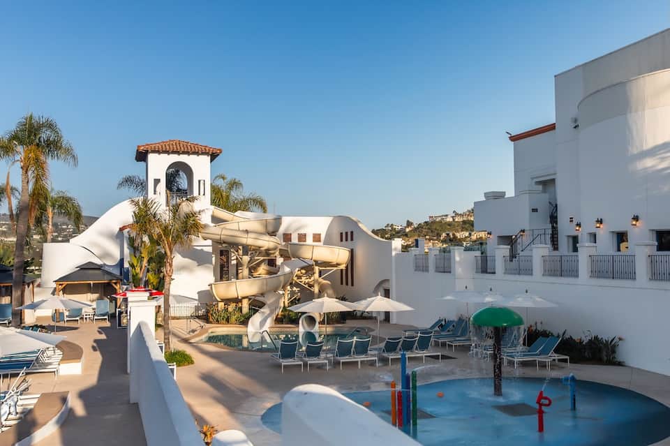 Resort courtyard with white architecture, water feature, lounge seating, palm trees, and splash pad