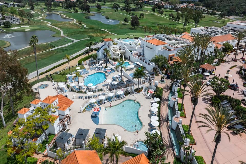 Aerial view of resort with multiple pools, orange-roofed buildings, golf course, and palm trees