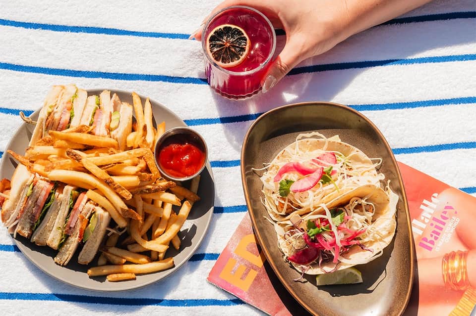 Overhead view of sandwich, fries, and fish tacos with red drink on striped beach towel