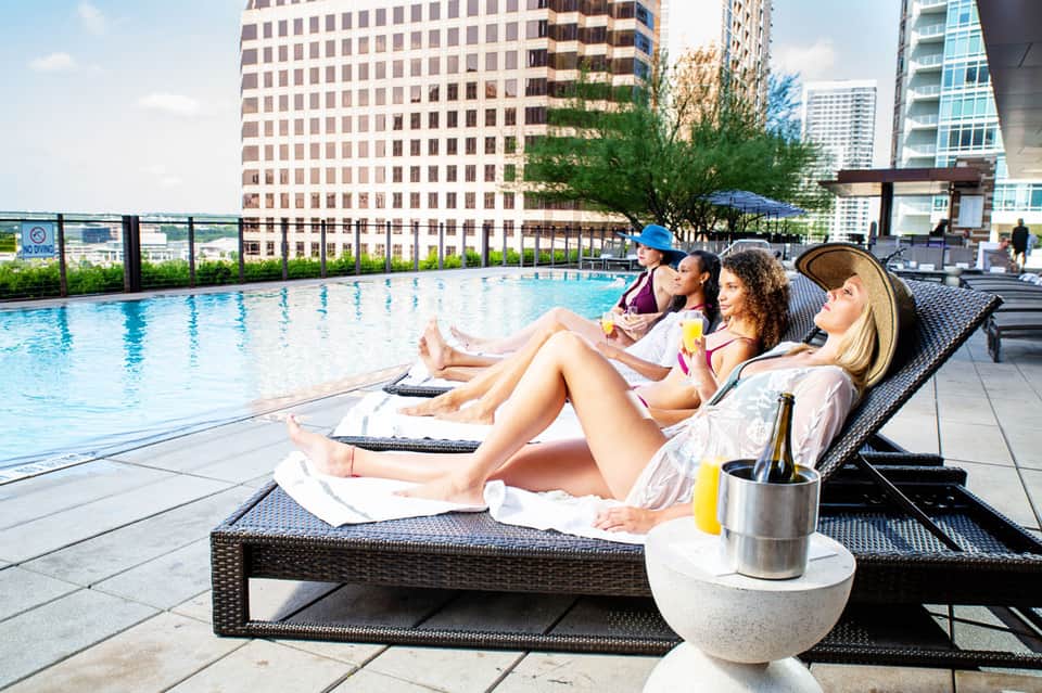 Four women relaxing on black lounge chairs by an urban rooftop pool with city skyline backdrop