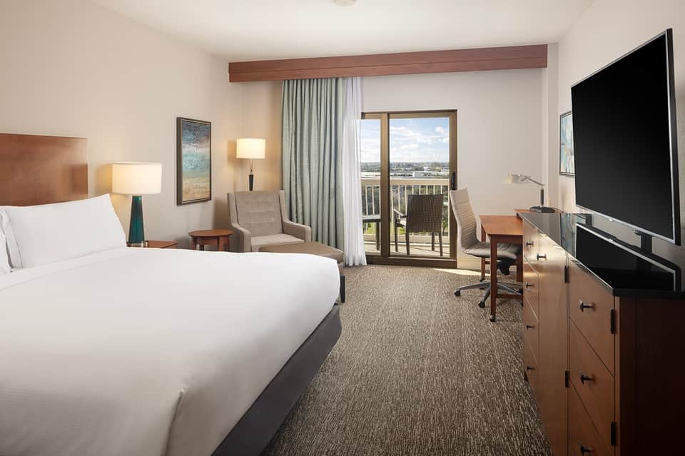 Hotel bedroom with queen bed, balcony overlooking waterfront, sitting area, TV, and modern furnishings
