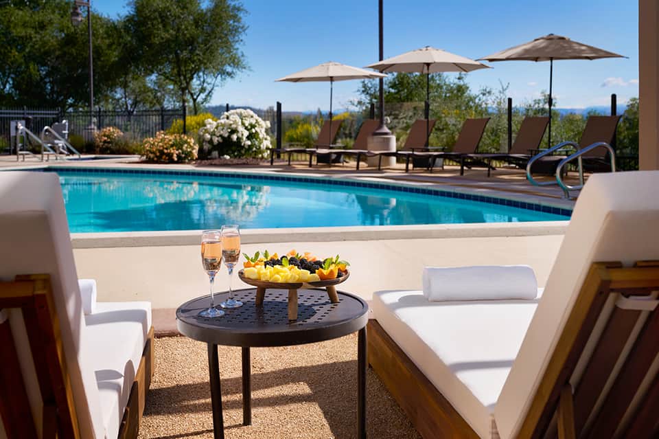 Resort pool with champagne glasses and fruit platter on lounge chairs overlooking mountains