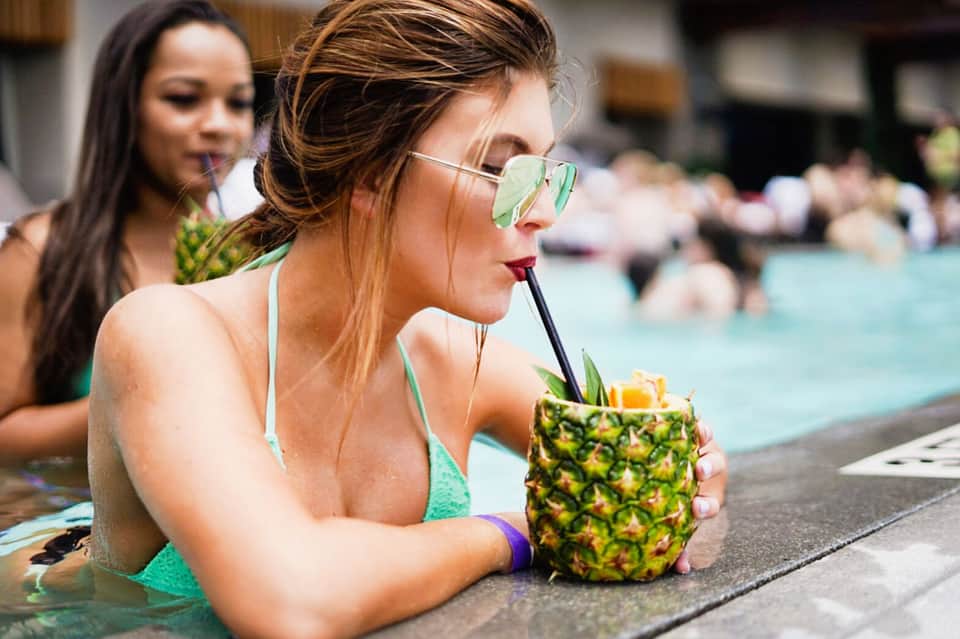 Woman in green bikini and sunglasses enjoying pineapple drink at pool edge with other guests in background