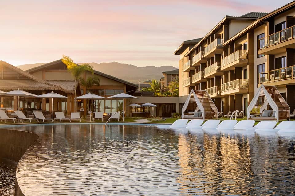 Luxury resort pool at sunset with mountain views, white cabanas, lounge chairs, and multi-story hotel building