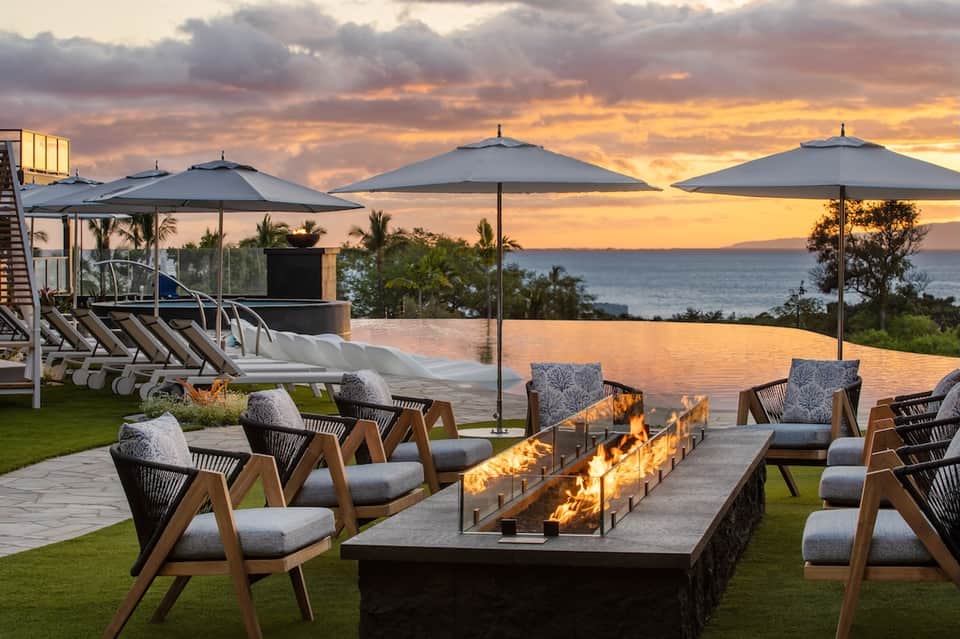 Oceanfront terrace at sunset with lounge chairs, umbrellas, fire pit, and coastal landscape views during golden hour