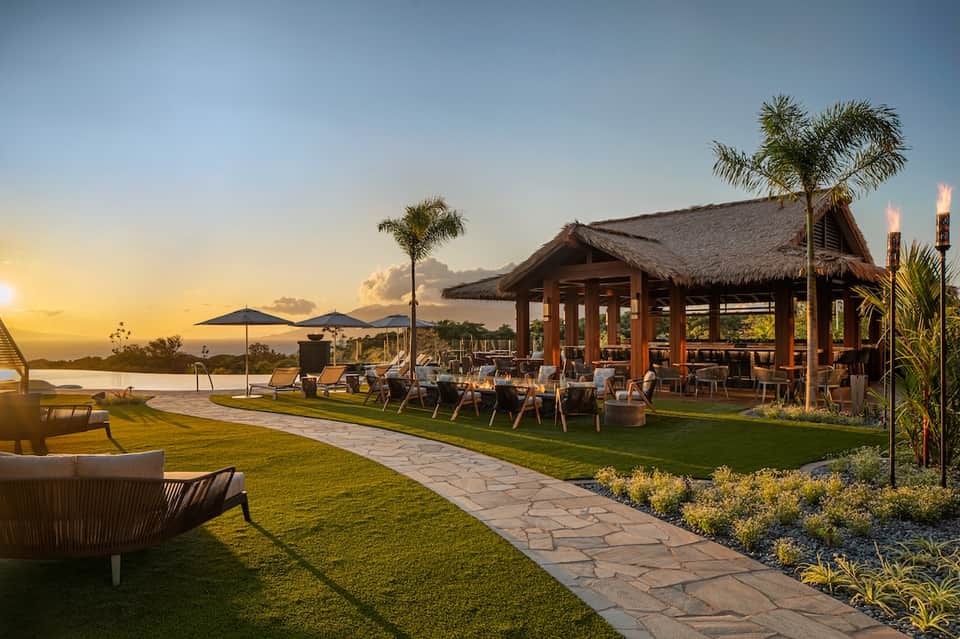 Tropical resort at golden sunset with thatched-roof pavilion, manicured lawn, palm trees, and infinity pool