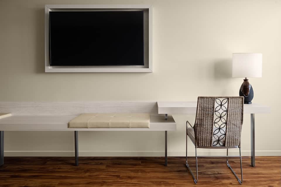 Modern hotel room seating area with beige bench, patterned chair, desk, lamp, and wall-mounted flat-screen TV