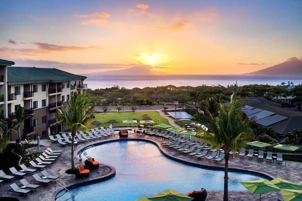 Scenic resort pool at sunset with mountain views, lounge chairs, palm trees, and curved infinity pool