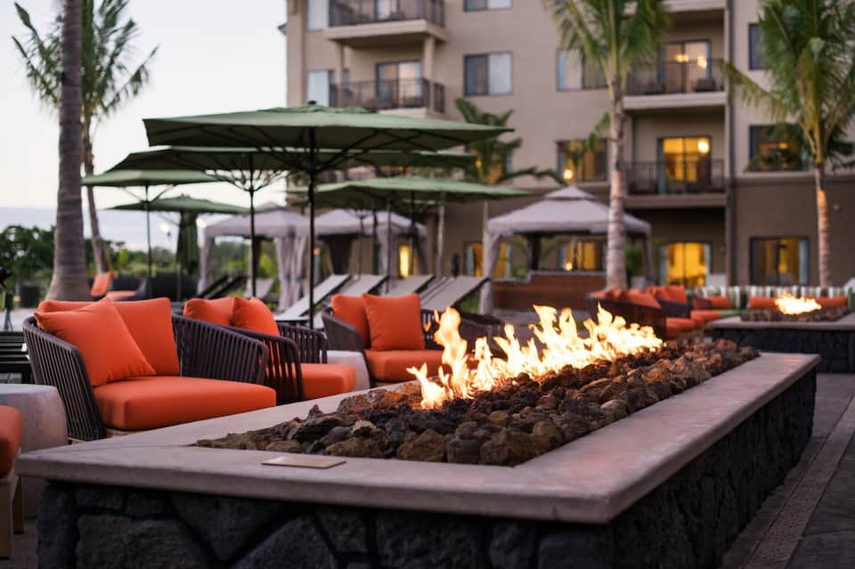 Resort courtyard at dusk with fire pit, orange lounge chairs, umbrellas, and palm trees