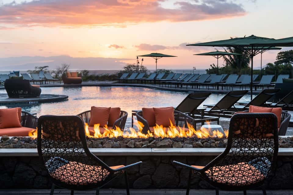 Sunset view of infinity pool with lounge chairs, fire pit, and architectural water features