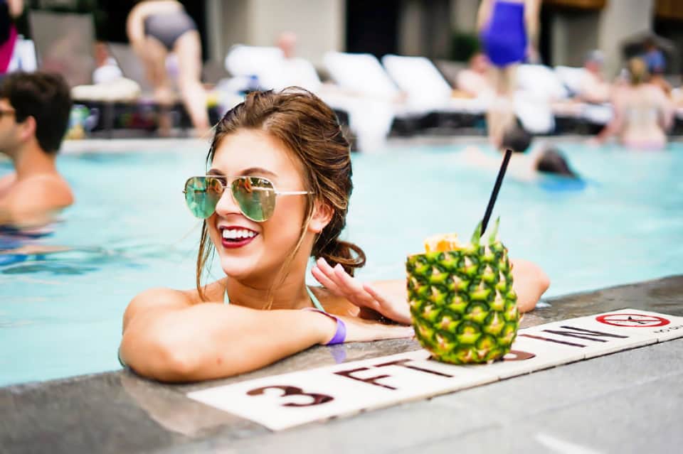 Woman in aviator sunglasses enjoying a pineapple drink at the edge of a crowded resort pool