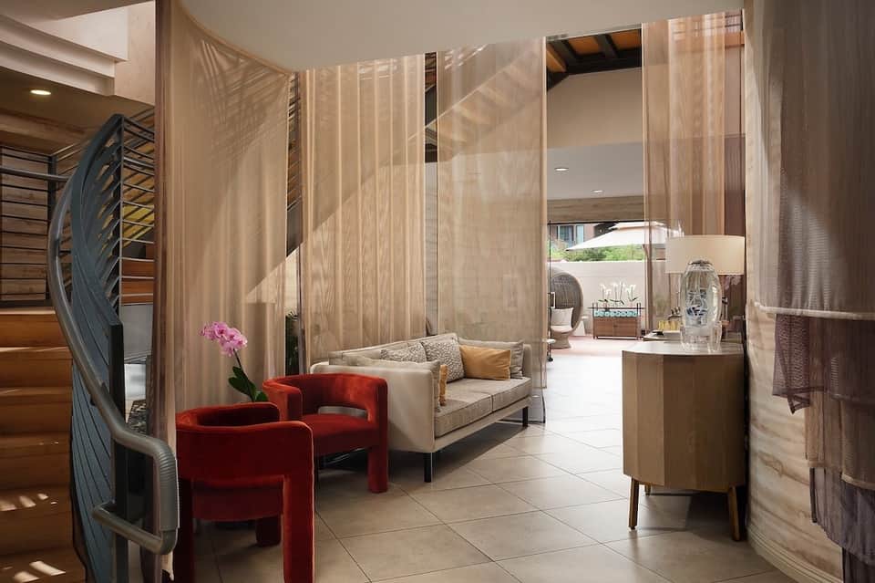 Luxury spa suite with sheer curtains, red accent chairs, beige sofa, wooden furnishings, and terraces