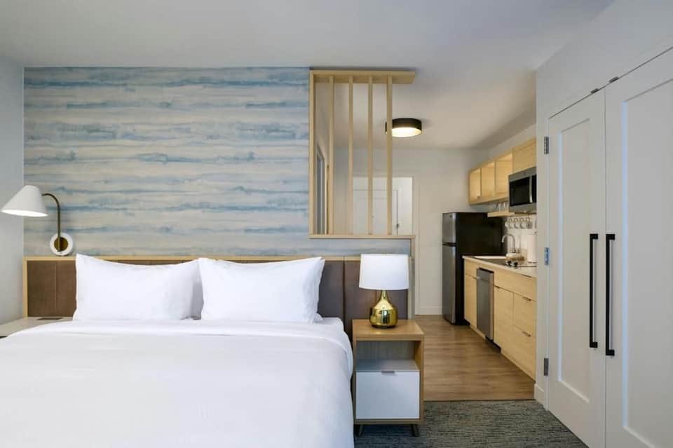 Compact hotel room with queen bed, kitchenette, wood accents, and blue wallpaper