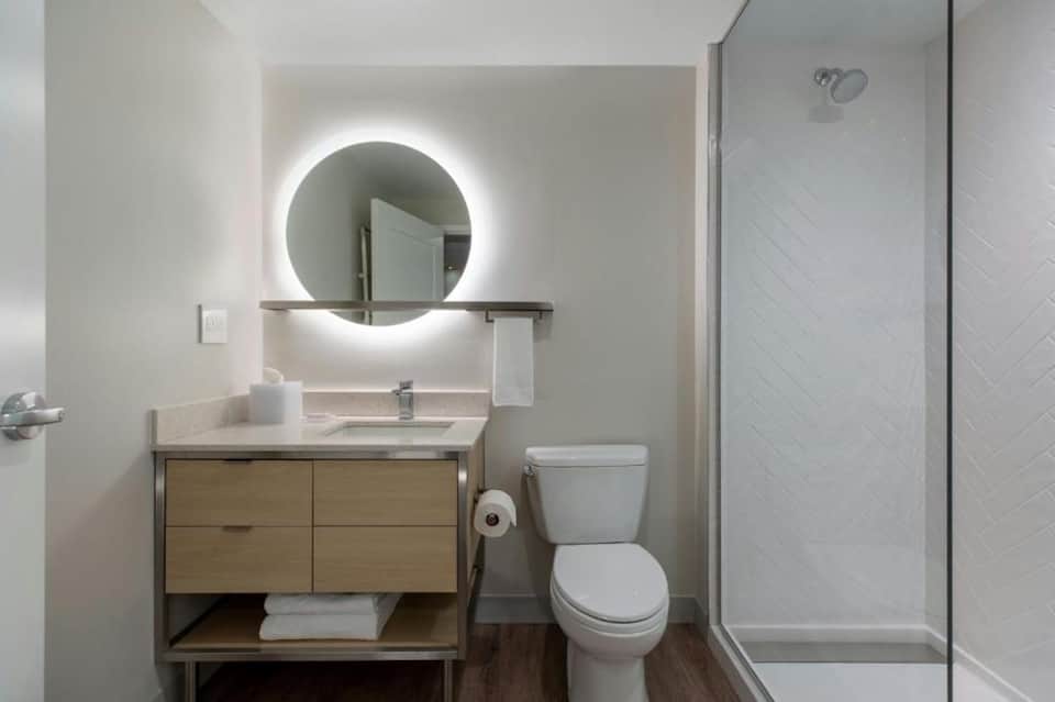 Contemporary bathroom with illuminated circular mirror, wooden vanity, and walk-in shower