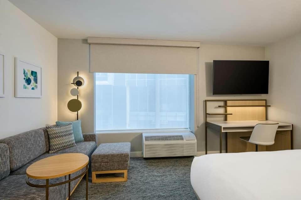 Hotel room with sofa, desk, TV, window with city view, and contemporary furnishings in neutral tones