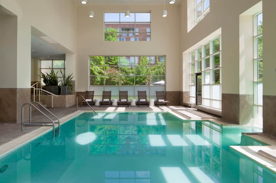 Indoor heated pool with floor-to-ceiling windows, lounge chairs, and natural lighting