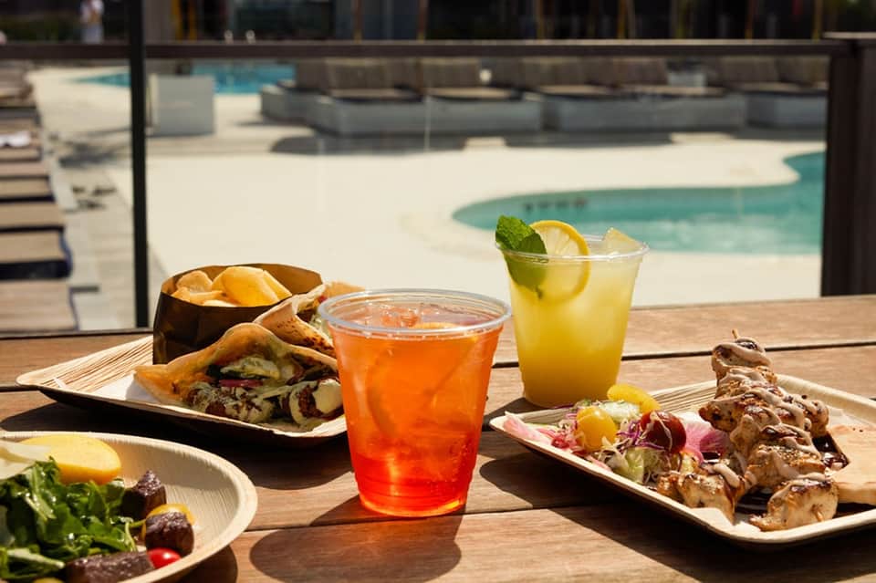 Poolside dining with cocktails, appetizers, and food plated on wooden table overlooking pool