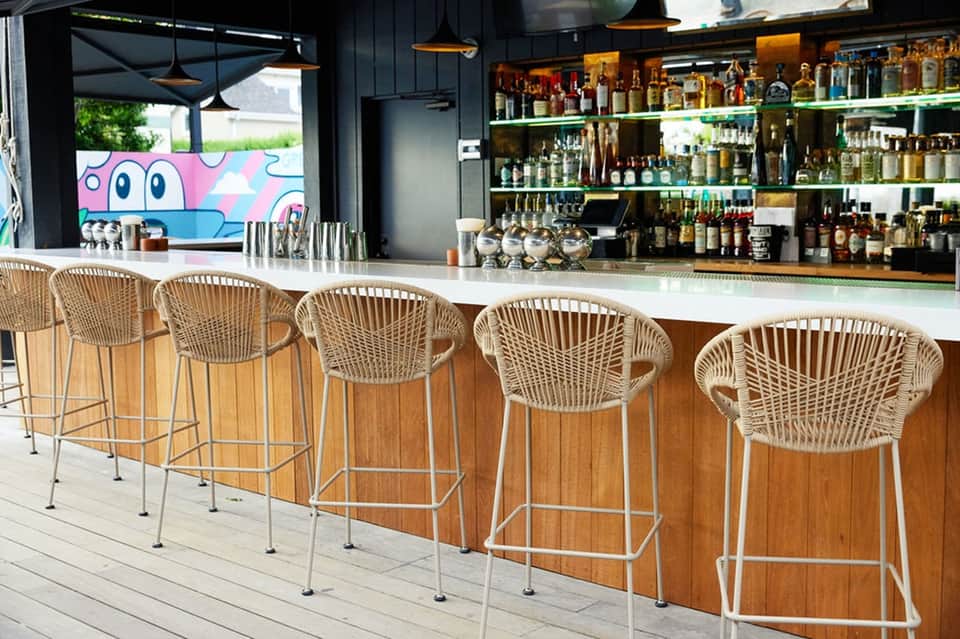 Modern beachfront bar with white woven stools, extensive liquor shelves, and colorful artwork
