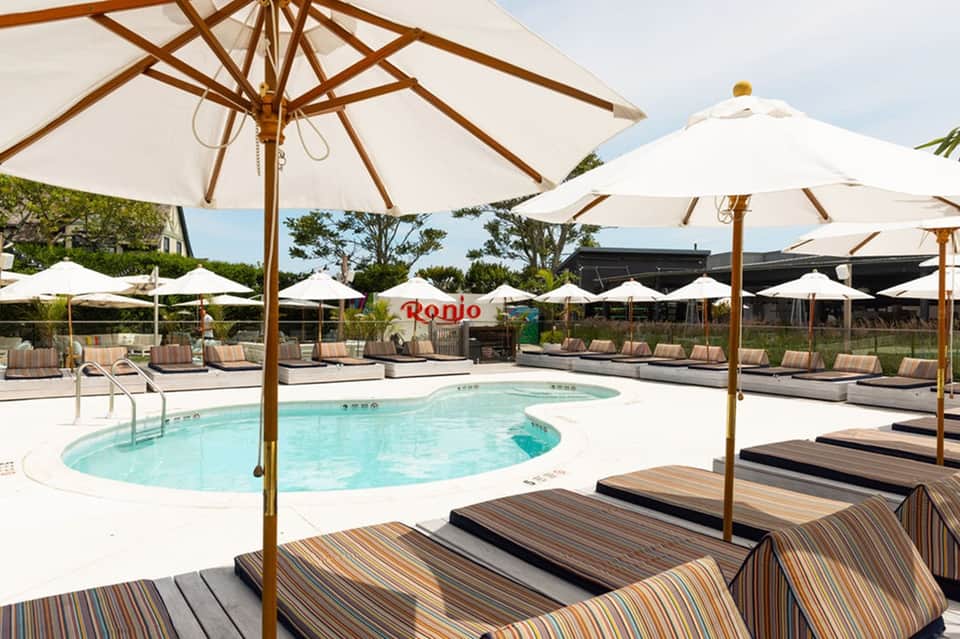 Resort pool area with lounge chairs, wooden umbrellas, white shade structures, and manicured landscaping