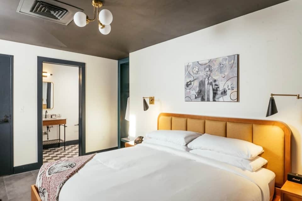Contemporary hotel bedroom with mustard-yellow headboard, ensuite bathroom, and modern lighting fixtures