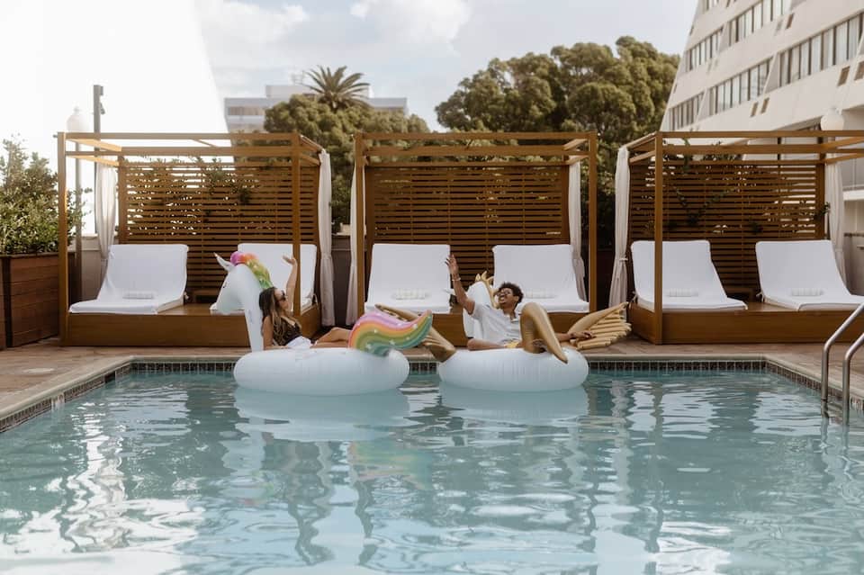 Guests relaxing in pool with private cabanas featuring white lounges and wooden pergola structures