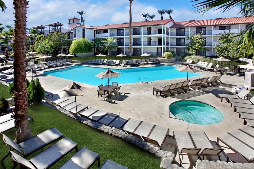 Resort pool area with bright turquoise water, lounge chairs, umbrellas, white building, and palm trees