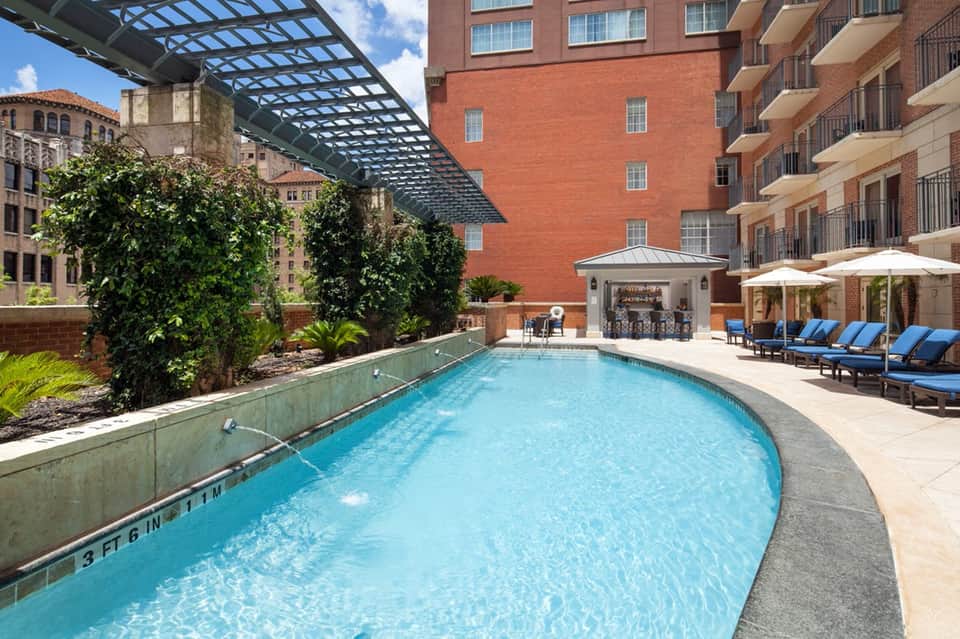 Urban rooftop pool with blue loungers, climbing ivy walls, and surrounding brick buildings