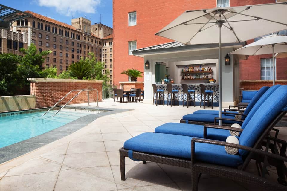 Urban rooftop pool with lounge chairs, bar area, umbrellas, and surrounding brick buildings