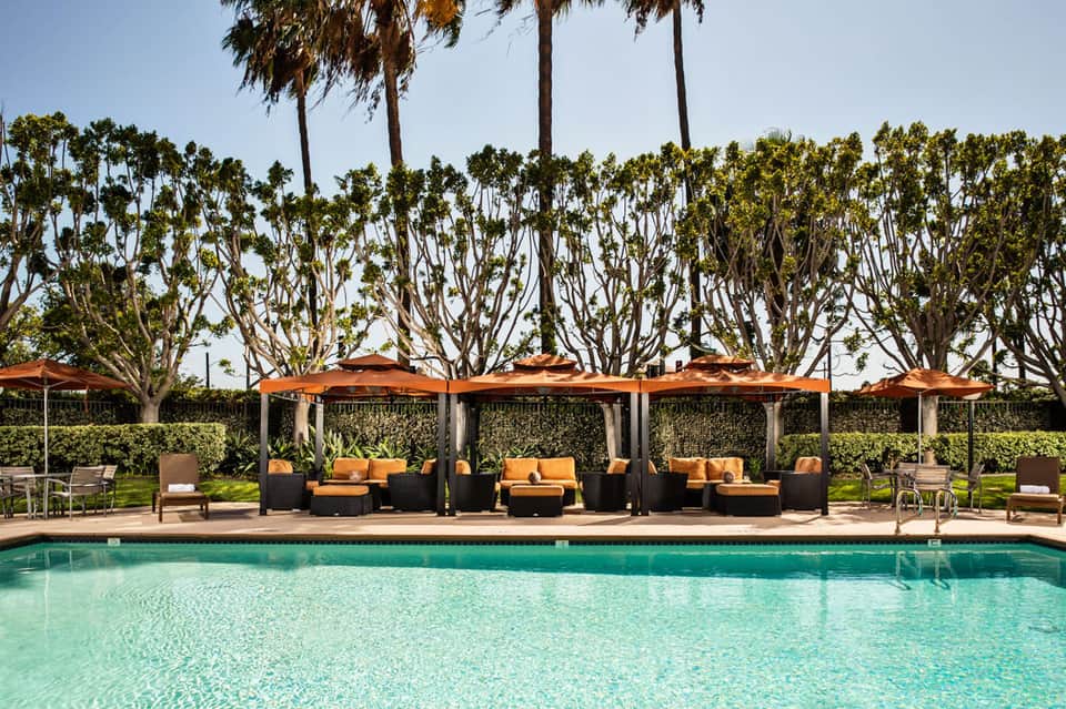 Resort pool with turquoise water, orange lounge seating under pergolas, tall palm trees, and manicured lawn