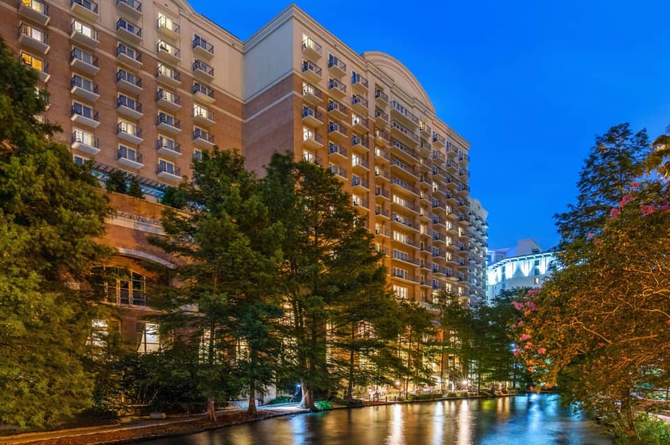 Brick residential building reflected in canal at dusk with illuminated windows and surrounding trees