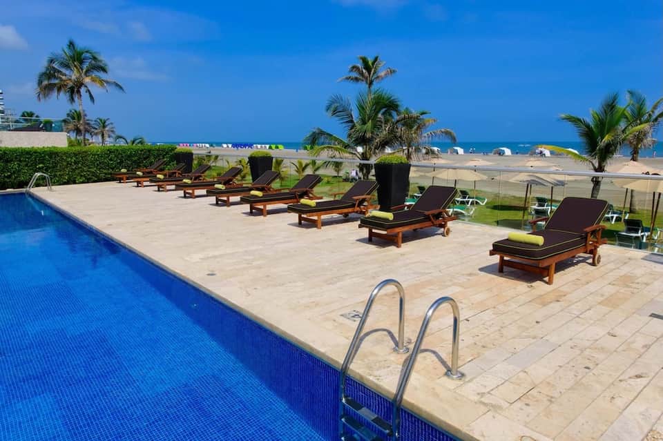 Beachfront pool with wooden lounge chairs, palm trees, and ocean view