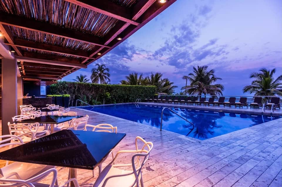 Illuminated resort pool at dusk with lounge chairs, covered seating area, and palm trees silhouetted against twilight sky