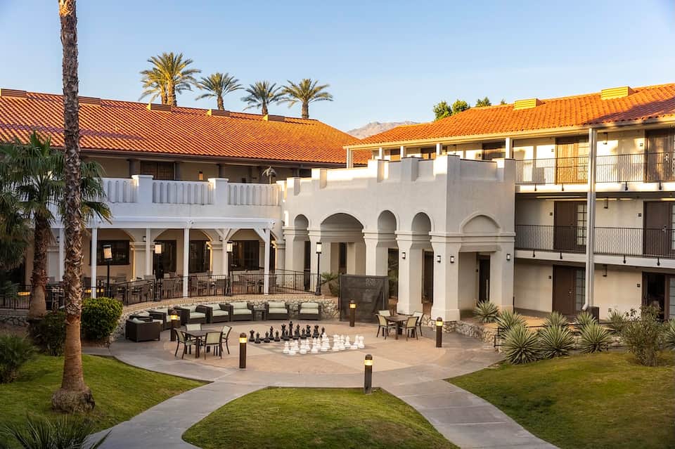Mediterranean-style courtyard with white columns, arched doorways, palm trees, and giant chessboard