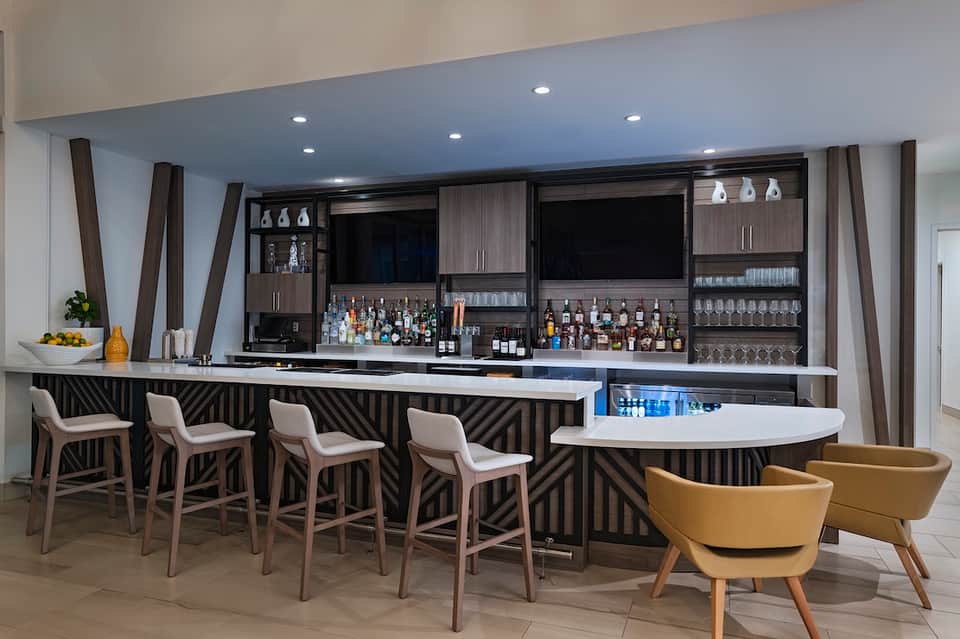 Modern hotel bar with dark geometric wood design, white bar seating, and liquor display