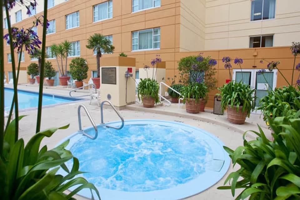 Hot tub surrounded by potted plants with resort building and larger pool visible in background courtyard