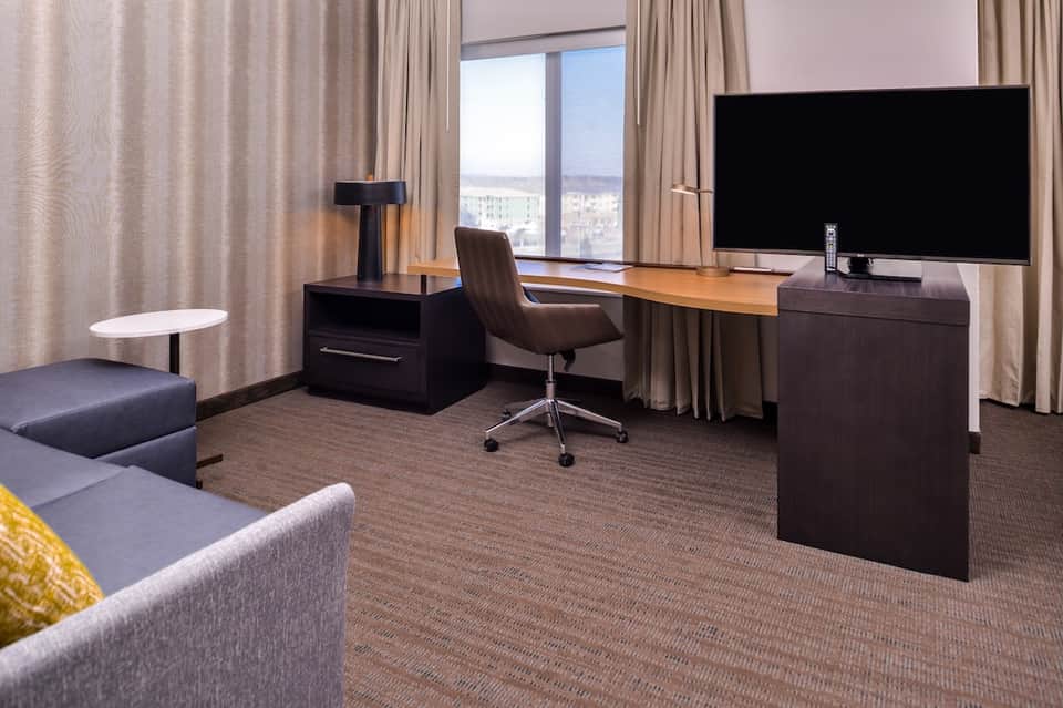 Hotel suite with dark wood desk, flat-screen TV, and city views through large windows with beige curtains