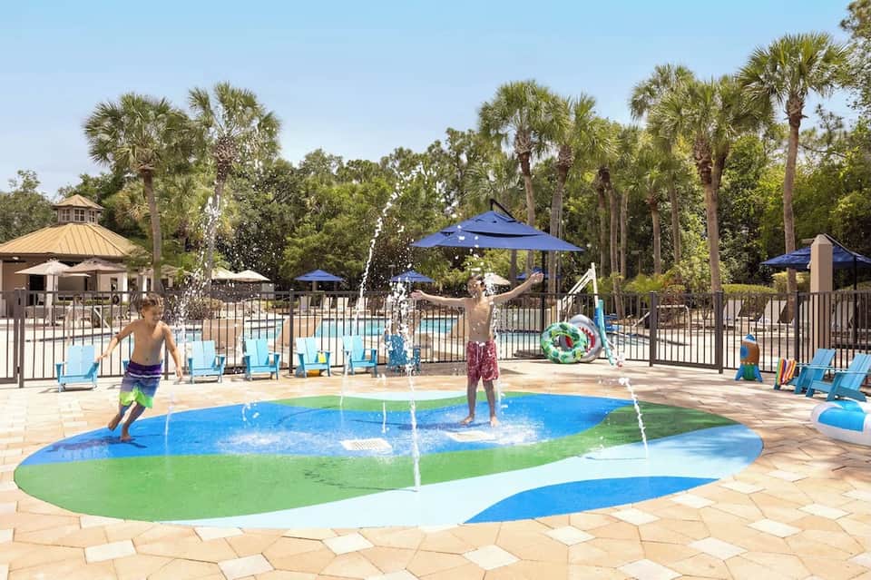 Family splash pad and pool area with colorful interactive water features surrounded by palm trees