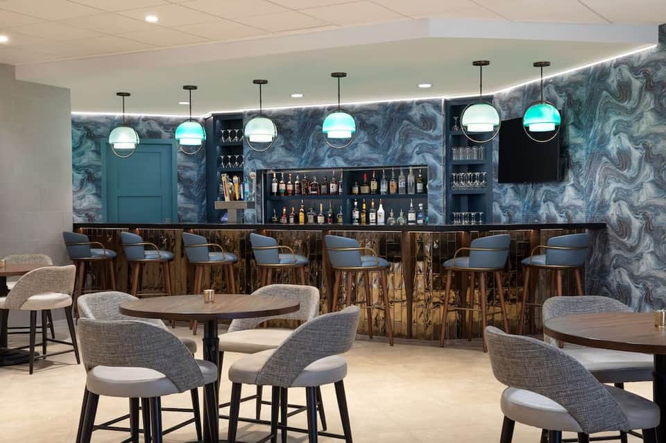 Contemporary hotel bar lounge with marble accent wall, wood bar seating, spherical pendant lights, and casual seating