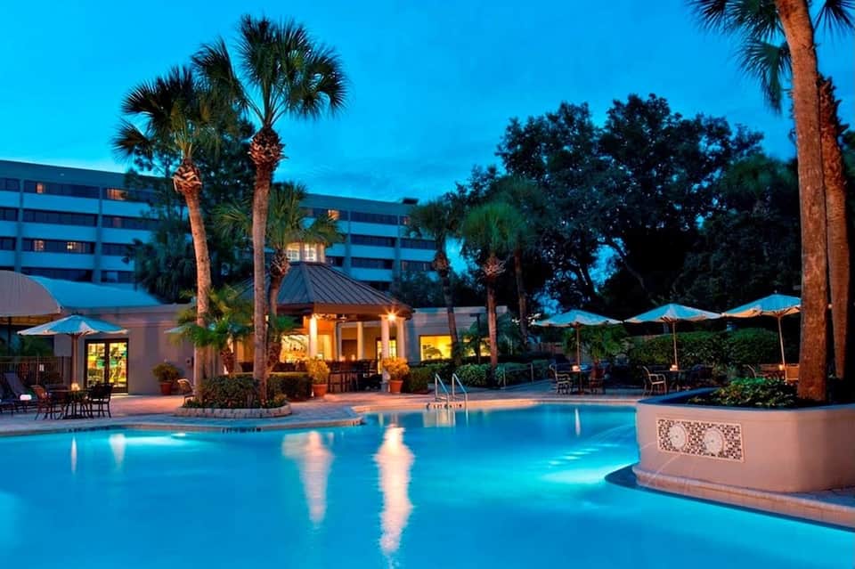 Resort pool at dusk with illuminated water, illuminated cabana, palm trees, umbrellas, and hotel building reflected in pool