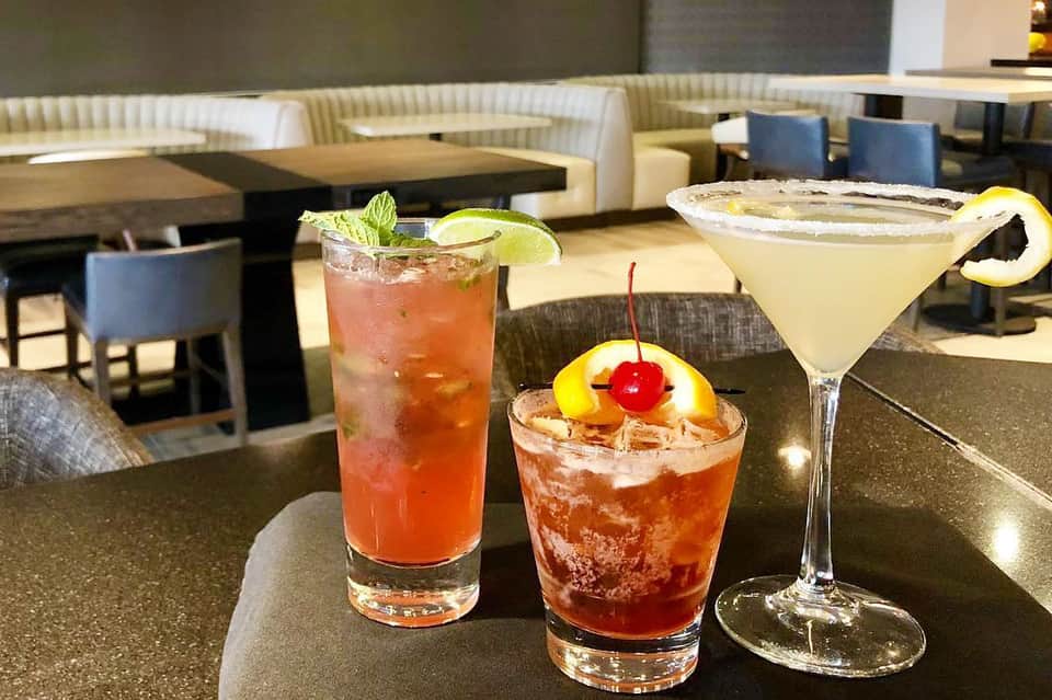 Three colorful cocktails on dark bar counter with lounge seating area in background