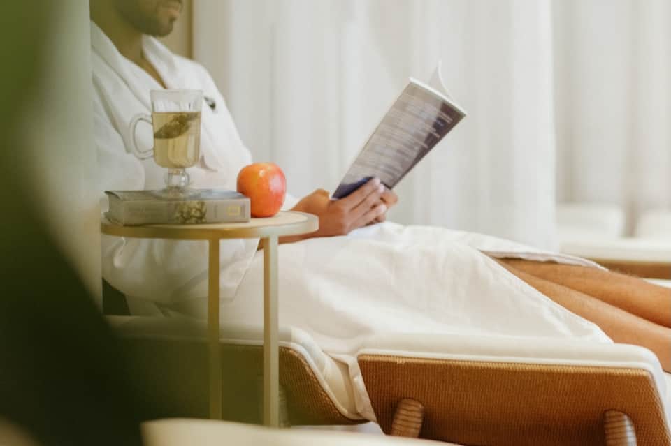 Woman in white robe reading on bed with tea, apple, and books on side table