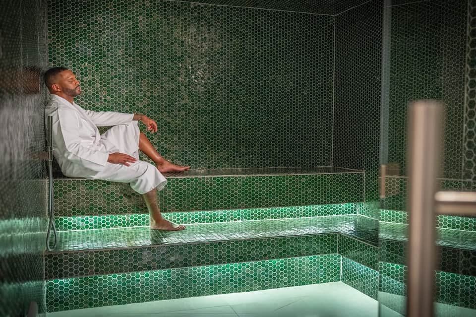 Man relaxing in hotel steam room with green hexagonal tile walls and bench seating.