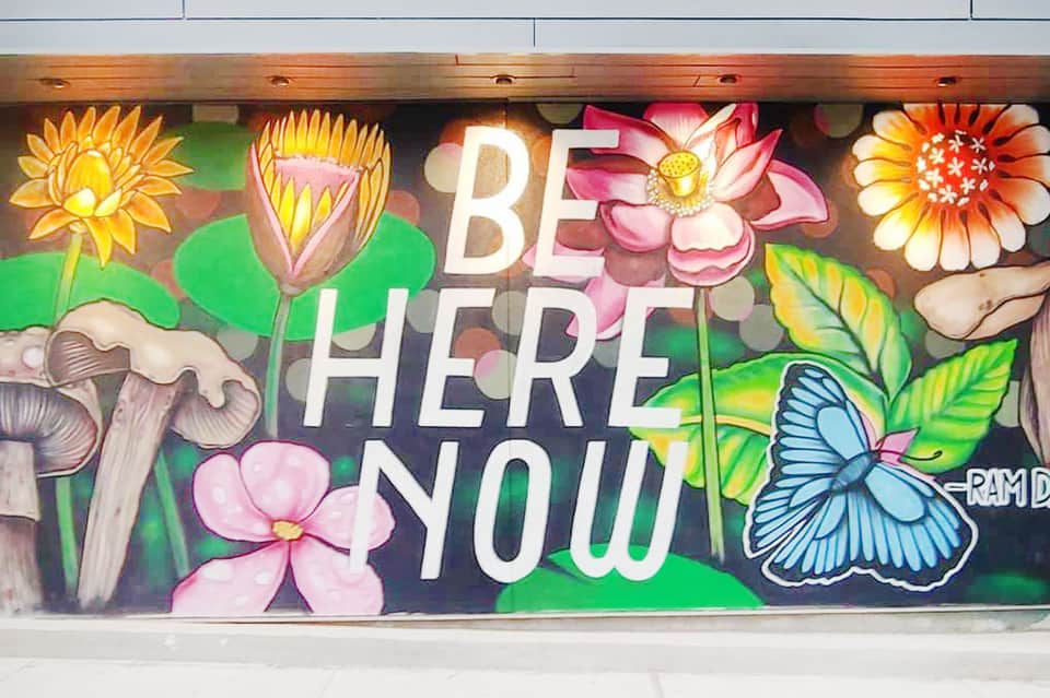 Colorful mural with flowers, mushrooms, and butterfly with 'BE HERE NOW' text
