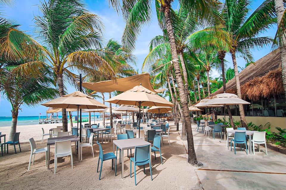 Beachfront dining area with shade umbrellas, white furniture, and palm trees on sand