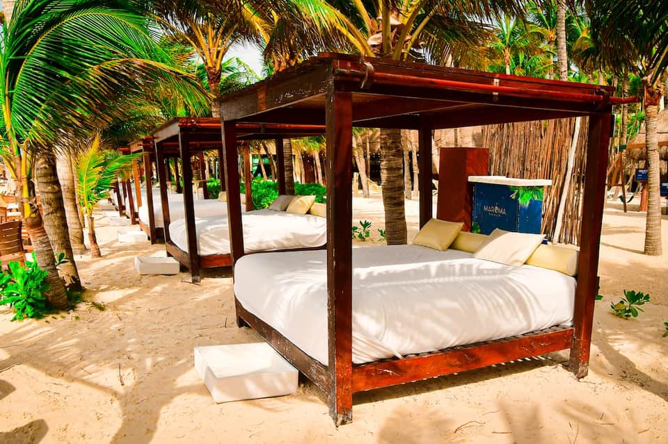 Beach cabana beds with dark wood frames, white bedding, and yellow pillows among palm trees on sandy shore