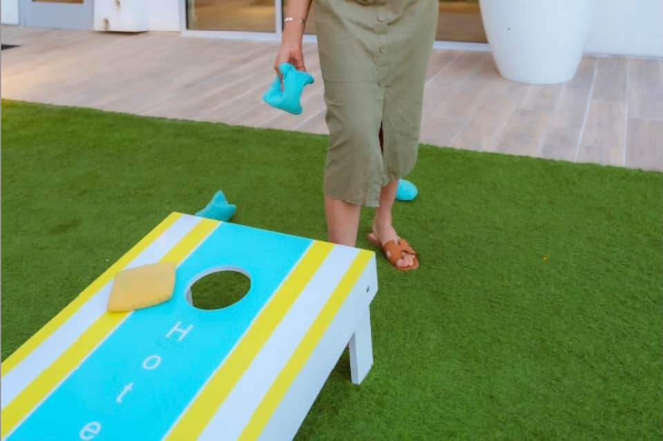 Person playing cornhole on artificial grass with blue and yellow game board